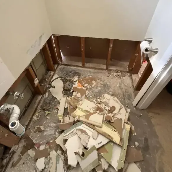Removed drywall and debris cover a bathroom floor beside exposed lower wall framing