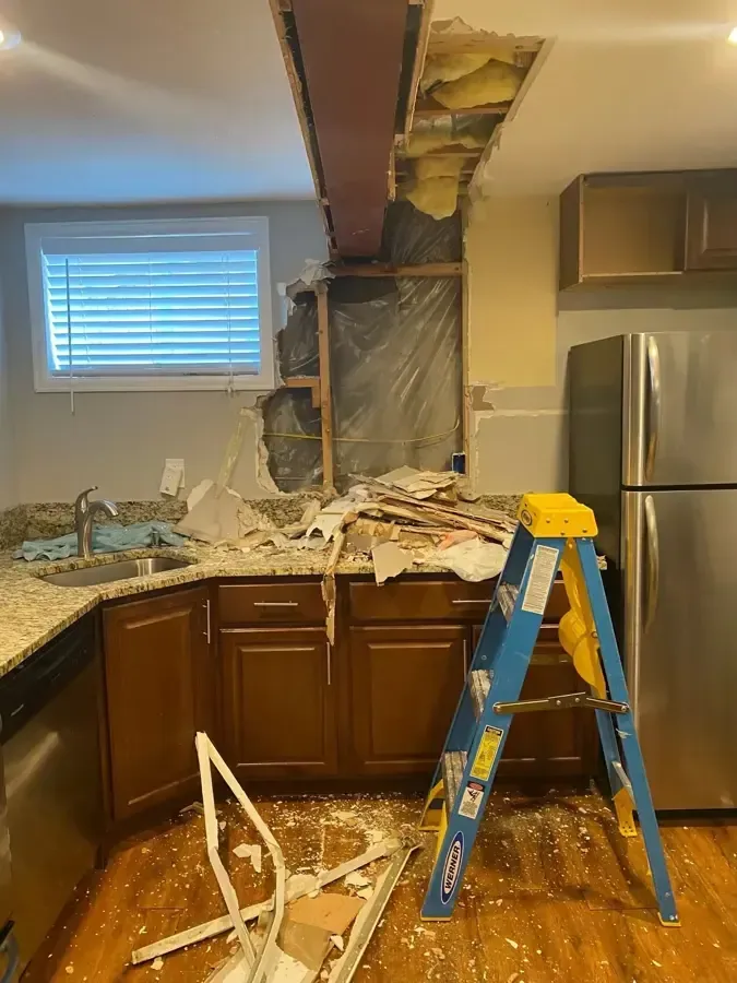 An opened kitchen ceiling exposes framing above a ladder and protected kitchen cabinets