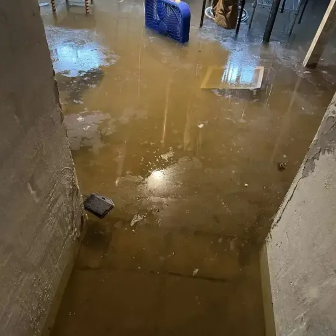 Muddy standing water covers a basement floor between exposed wall bases and belongings