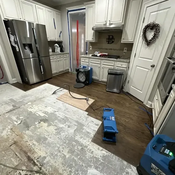 A dehumidifier and blue air movers stand on partially exposed flooring inside a kitchen