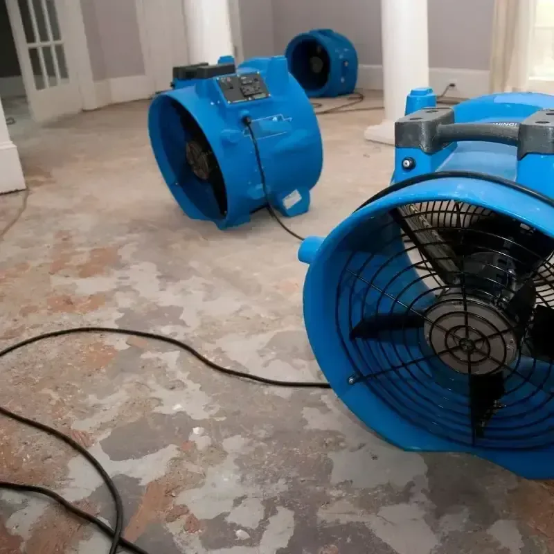 Blue air movers stand on an exposed floor beside patched walls during drying