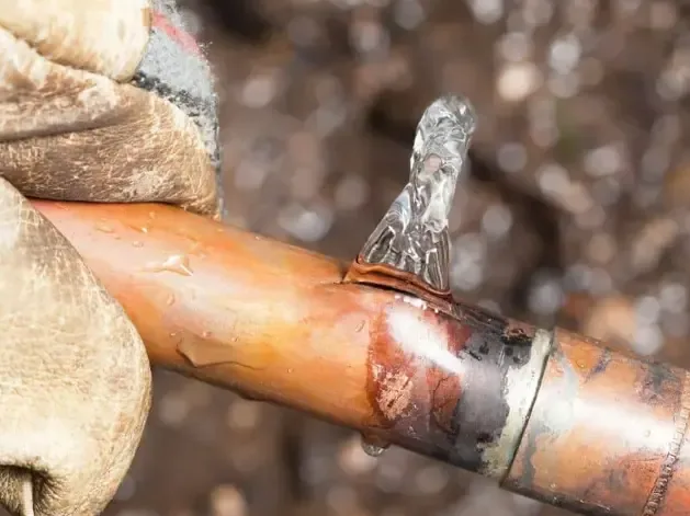 A gloved hand holds a copper pipe with water spraying from its split