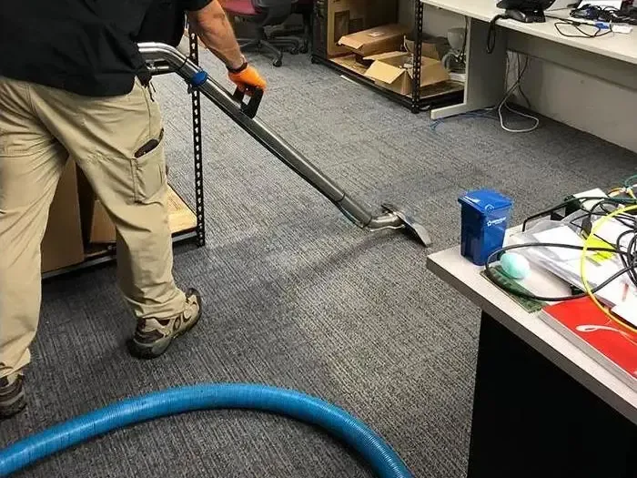 A technician uses an extraction wand on office carpet beside desks and equipment