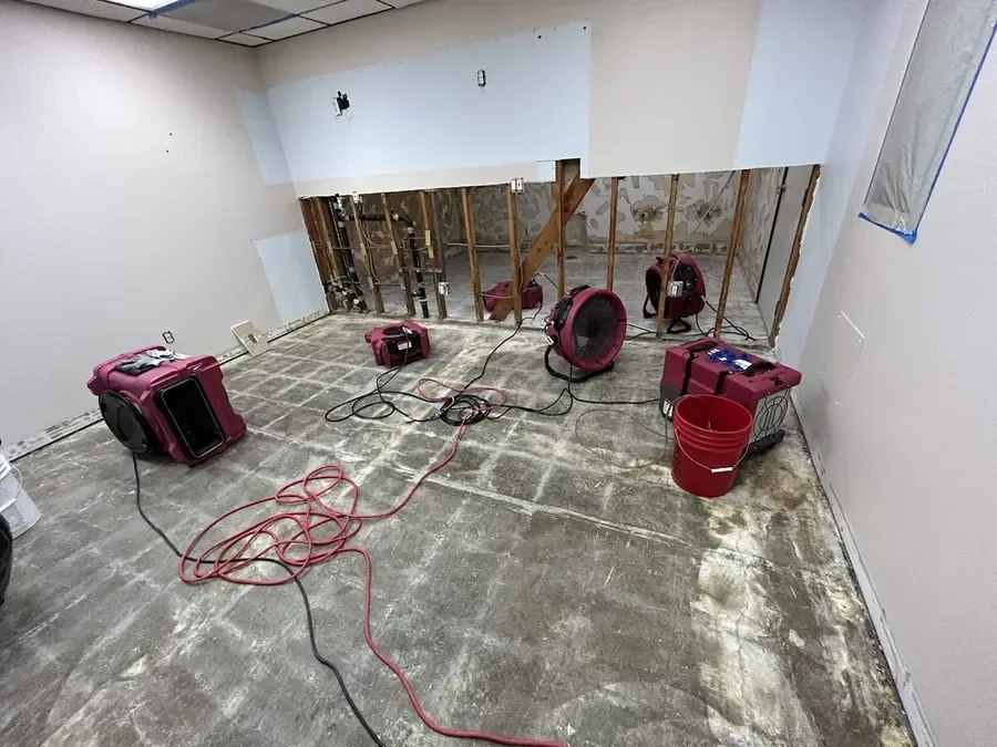 Air movers and dehumidifiers dry exposed concrete flooring beside opened lower wall cavities
